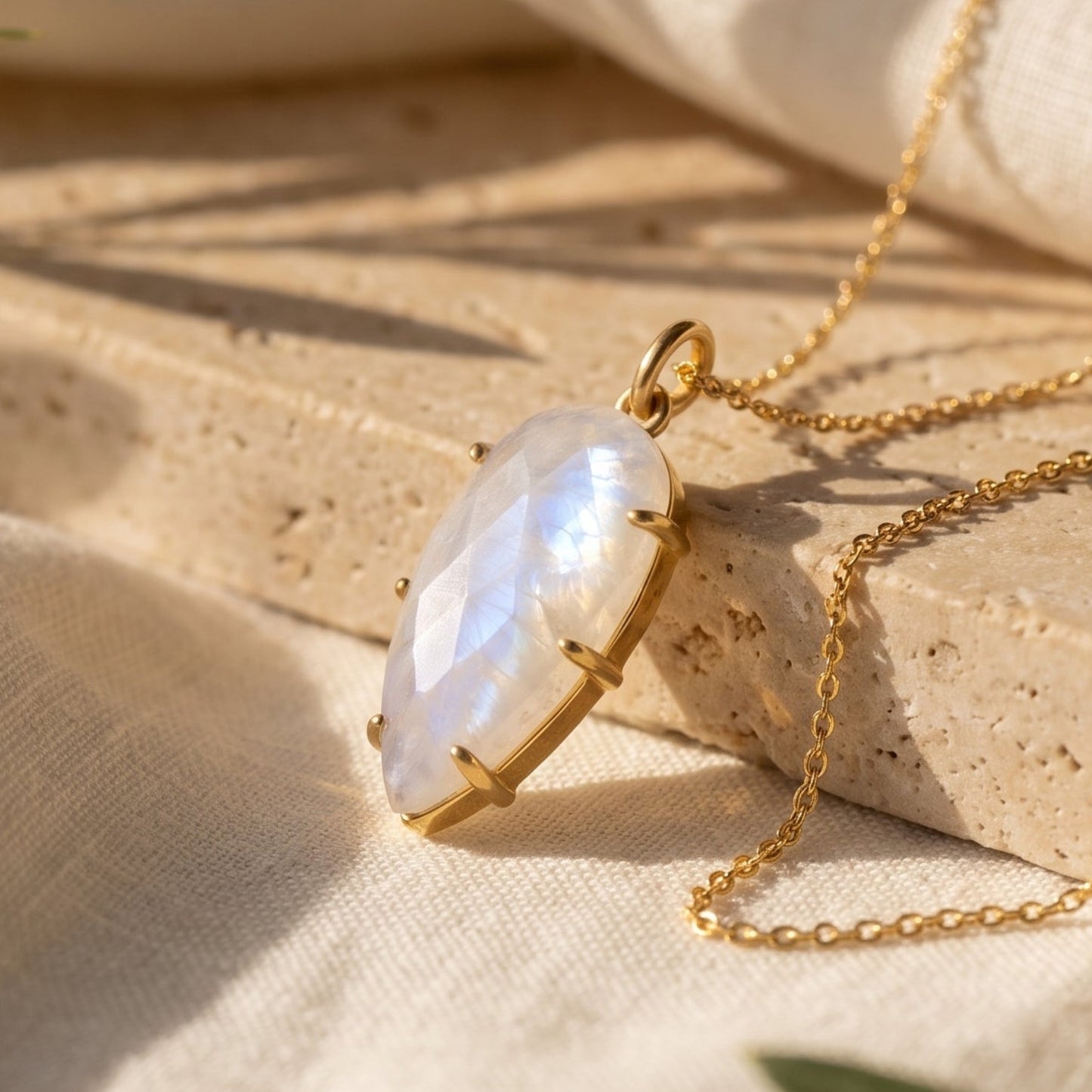 Gold necklace with a marquise-shaped iridescent moonstone pendant, styled on draped cream linen fabric and a stone slab with olive leaves.
